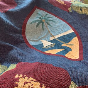 May include: A blue and red woven tapestry featuring the flag of Samoa. The flag depicts a white sailboat on a blue ocean with a palm tree on a sandy beach.