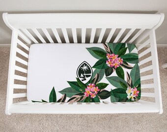 Op de afbeelding: Witte wieg met een hoeslaken met een tropisch bloemenontwerp. Het laken heeft een witte achtergrond met groene bladeren, roze bloemen en een zwart-wit embleem. De tekst "ISLAND LIFE" staat rechtsonder.