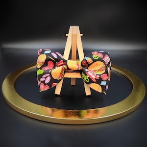 May include: A black bow tie with a colorful print of various fast food items, including hamburgers, french fries, and hot dogs. The bow tie is displayed on a wooden easel.