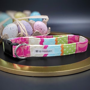 May include: A colorful patchwork dog collar with a pink, green, blue, and white pattern. The collar has a black buckle and a silver D-ring. The collar is on a gold-colored round surface. The text "R&R Haberdashery" is on the collar.