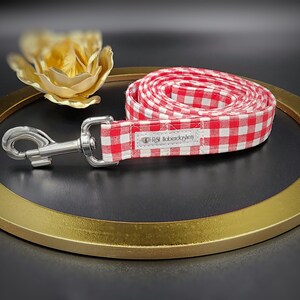 May include: A red and white gingham dog leash with a silver metal clasp. The leash is coiled on a gold-colored circular tray. The label on the leash reads "R&M Laberdashery".