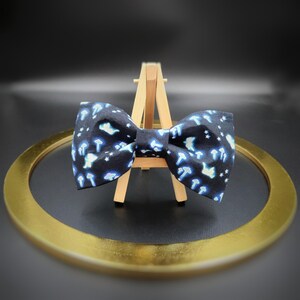 May include: A dark blue bow tie with a pattern of white and blue mushrooms and stars. The bow tie is displayed on a small wooden easel, with a gold-colored ring in the foreground.