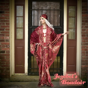 Pode incluir: Uma mulher em um conjunto bordô, incluindo um robe com estampas florais douradas, um adorno de cabeça combinando e lingerie. A roupa é adornada com joias douradas. A imagem é tirada em frente a um edifício de tijolos com uma porta.