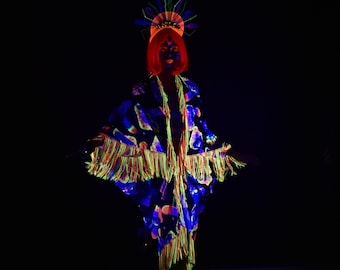 May include: A person wearing a blue and black patterned robe with yellow fringe and a neon green and orange headpiece. The person is standing in a dark room with a neon sign that says "Jezebel's Boudoir" in the bottom right corner.