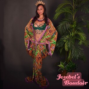 Può includere: Una donna indossa una corona d'oro e una lunga veste fluida con stampa leopardata e design a foglie tropicali. La veste ha una cintura rosa e bordi perlati. Indossa anche un reggiseno di pizzo verde e una collana con gemme rosse e blu. Lo sfondo è una parete grigia scura con una pianta in vaso.
