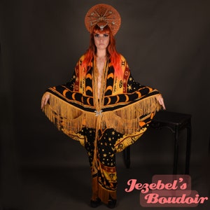 May include: A woman wearing a black and gold fringed robe with a celestial print. The robe features a crescent moon, stars, and zodiac symbols. She is wearing a crown with rhinestones and has red hair. The text "Jezebel's Boudoir" is visible in the bottom right corner of the image.