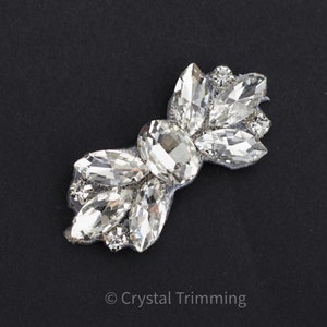 May include: A clear crystal embellishment with a large oval-shaped crystal in the center and smaller, teardrop-shaped crystals surrounding it. The embellishment is attached to a silver mesh backing.