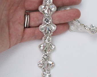 May include: A close-up of a crystal beaded trim with teardrop-shaped crystals. The trim is on a white background.