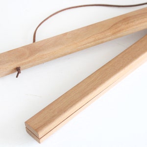 May include: Wooden poster hanger with a natural finish. The two wooden bars are connected by a brown leather cord. The top bar has a small leather loop. The wood has a light brown colour.