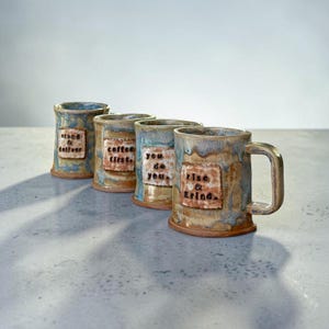 May include: A set of four ceramic mugs with a mottled blue and brown glaze. Each mug features a rectangular patch with stamped black text. The text reads: stand & deliver, coffee first, you do you, and rise & grind. Each mug has a sturdy handle.