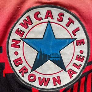 May include: Close-up of a red and black jersey with a circular patch. The patch features a blue star with a cityscape silhouette, surrounded by the words "NEWCASTLE BROWN ALE" in red. The jersey has a gradient from red to orange.