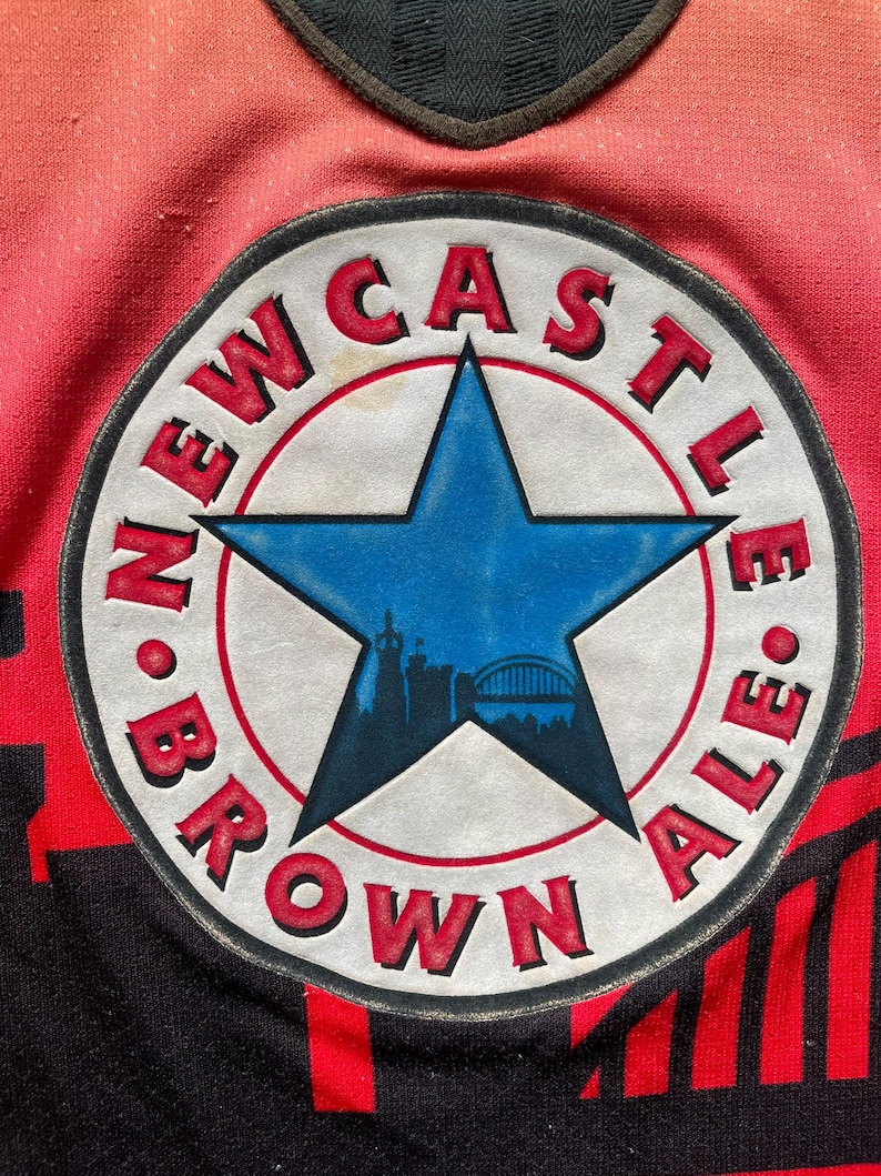 May include: Close-up of a red and black jersey with a circular patch. The patch features a blue star with a cityscape silhouette, surrounded by the words "NEWCASTLE BROWN ALE" in red. The jersey has a gradient from red to orange.