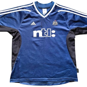 Op de afbeelding: Een blauw voetbaltenue met witte en zwarte accenten. Het tenue heeft het Adidas-logo, het Newcastle United-wapen en de tekst "ntl:" op de voorkant.