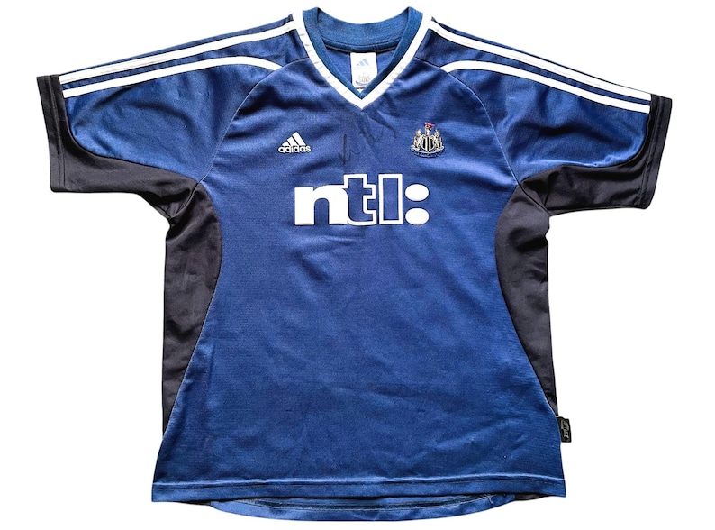 Op de afbeelding: Een blauw voetbaltenue met witte en zwarte accenten. Het tenue heeft het Adidas-logo, het Newcastle United-wapen en de tekst "ntl:" op de voorkant.