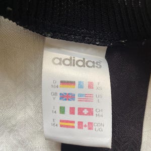 May include: Close-up of a white Adidas tag with black trim. The tag displays the Adidas logo and various country size charts, including US L and XS. The tag also shows the size 164.