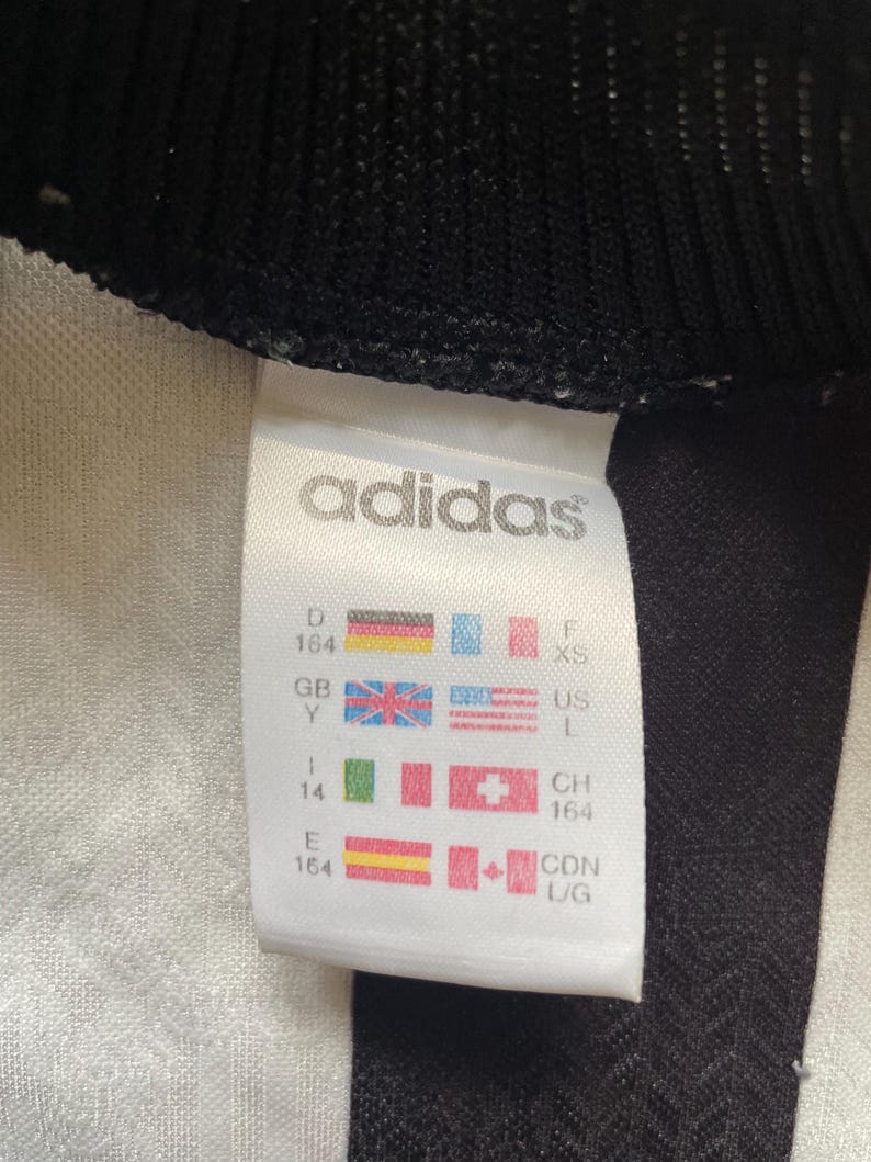 May include: Close-up of a white Adidas tag with black trim. The tag displays the Adidas logo and various country size charts, including US L and XS. The tag also shows the size 164.