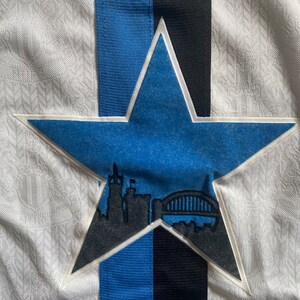 May include: A white, blue and black star-shaped patch with a silhouette of a city skyline and a bridge inside. The star is sewn onto a white fabric background.