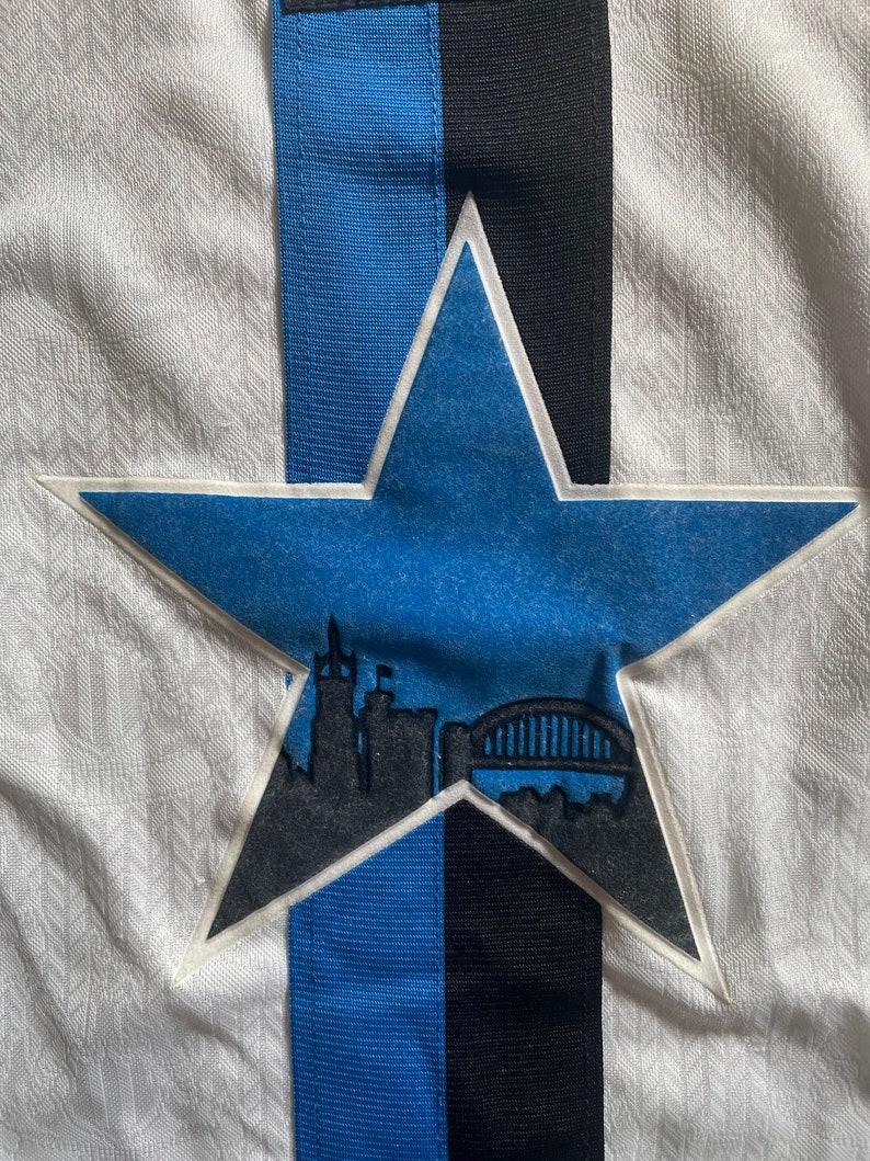 May include: A white, blue and black star-shaped patch with a silhouette of a city skyline and a bridge inside. The star is sewn onto a white fabric background.