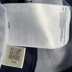 May include: Close-up of a white tag with the text "BLONE UPORABLJAITE MEHČALCA/PERITE IN LIKAJTE OBRNJENO" and "AHJ001". The tag is attached to a blue and gray fabric. A smaller tag with a barcode and the number "00606" is also visible.