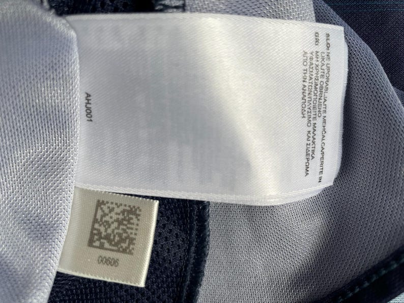May include: Close-up of a white tag with the text "BLONE UPORABLJAITE MEHČALCA/PERITE IN LIKAJTE OBRNJENO" and "AHJ001". The tag is attached to a blue and gray fabric. A smaller tag with a barcode and the number "00606" is also visible.
