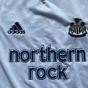 May include: White football shirt with a black Adidas logo, a Newcastle United crest, and the words "northern rock" in navy blue.