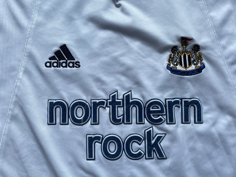 May include: White football shirt with a black Adidas logo, a Newcastle United crest, and the words "northern rock" in navy blue.