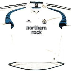 May include: White football shirt with black and blue accents. The shirt has the Adidas logo, the Newcastle United crest, and the text "northern rock" on the front.