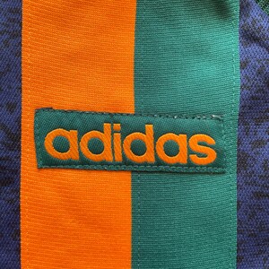 May include: Close-up of a section of athletic apparel. The design features vertical stripes in orange, teal, and navy blue. The Adidas logo is prominently displayed in orange on a teal patch.