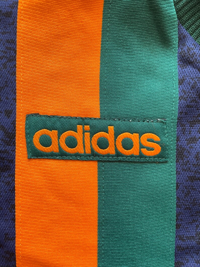 May include: Close-up of a section of athletic apparel. The design features vertical stripes in orange, teal, and navy blue. The Adidas logo is prominently displayed in orange on a teal patch.