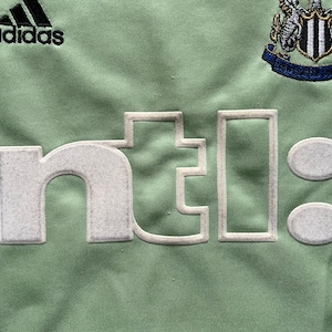 Pu&ograve; includere: Primo piano di un tessuto verde chiaro con il logo Adidas e uno stemma della squadra. Il testo "ntl:" &egrave; visualizzato in bianco sporco strutturato. Il design suggerisce una maglia sportiva o abbigliamento.