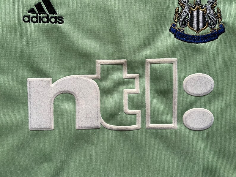 Pu&ograve; includere: Primo piano di un tessuto verde chiaro con il logo Adidas e uno stemma della squadra. Il testo "ntl:" &egrave; visualizzato in bianco sporco strutturato. Il design suggerisce una maglia sportiva o abbigliamento.