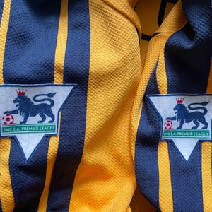 May include: Two yellow and blue striped soccer jerseys with a white patch on each side that says "The F.A. Premier League" and features a lion with a crown and a soccer ball.
