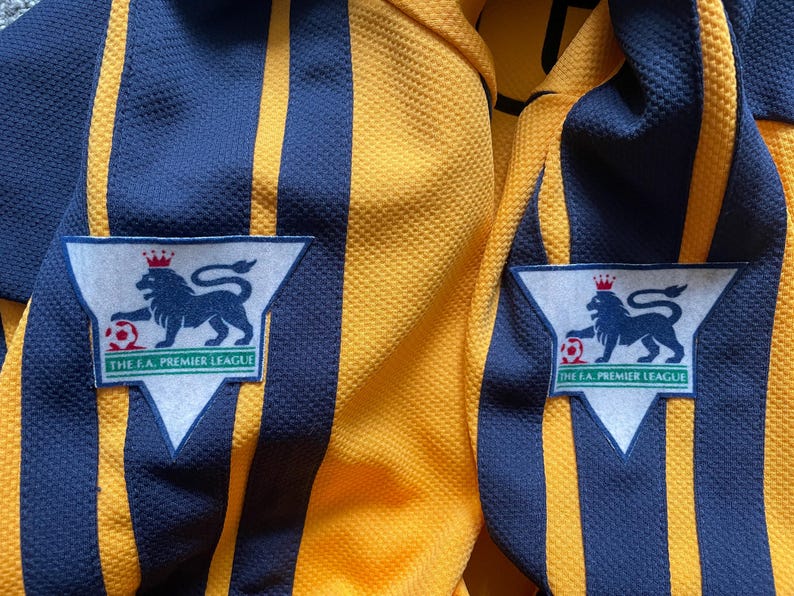 May include: Two yellow and blue striped soccer jerseys with a white patch on each side that says "The F.A. Premier League" and features a lion with a crown and a soccer ball.