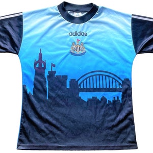 K&ouml;nnte beinhalten: Ein blaues und schwarzes Fu&szlig;balltrikot mit dem Adidas-Logo und dem Wappen von Newcastle United. Das Trikot zeigt eine Skyline von Newcastle, England, einschlie&szlig;lich der Tyne Bridge.