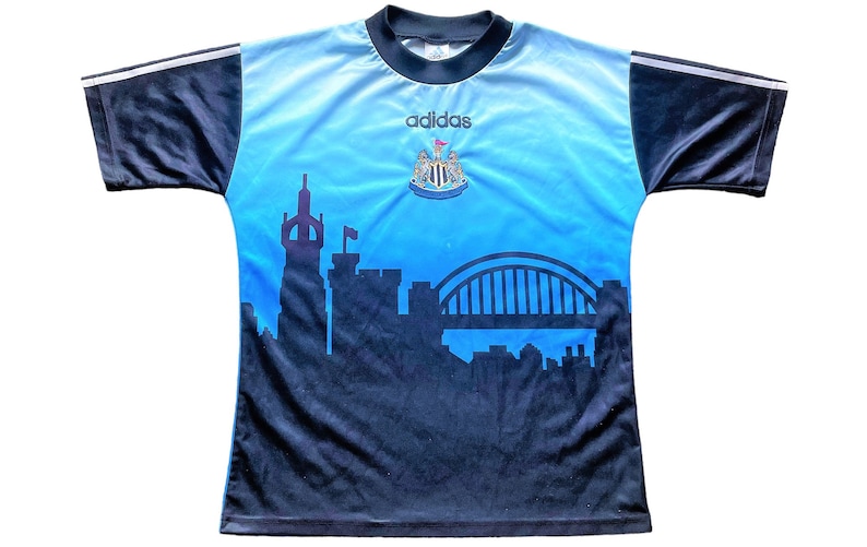 K&ouml;nnte beinhalten: Ein blaues und schwarzes Fu&szlig;balltrikot mit dem Adidas-Logo und dem Wappen von Newcastle United. Das Trikot zeigt eine Skyline von Newcastle, England, einschlie&szlig;lich der Tyne Bridge.