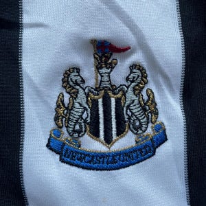 May include: A white and black striped soccer jersey with a blue and gold embroidered crest featuring a crown, two seahorses, and the text "NEWCASTLE UNITED".