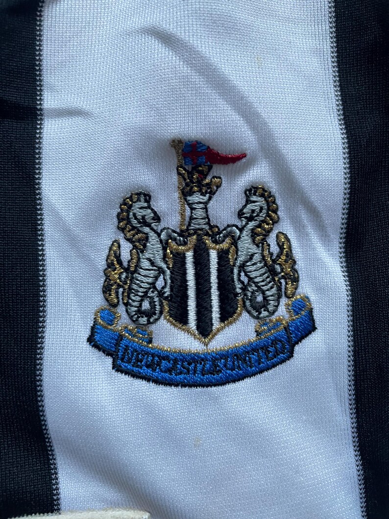 May include: A white and black striped soccer jersey with a blue and gold embroidered crest featuring a crown, two seahorses, and the text "NEWCASTLE UNITED".