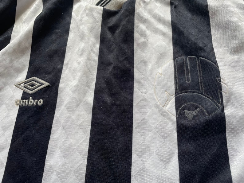 May include: Black and white striped soccer jersey with the logo "Umbro" and a team logo on the right side.
