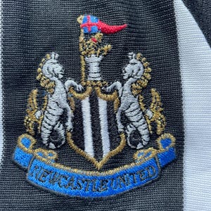 May include: Black and white striped soccer jersey with a black embroidered crest featuring a shield with black and white stripes, two white horses, and a gold crown. The crest is surrounded by a blue ribbon with the text "NEWCASTLE UNITED" in gold.