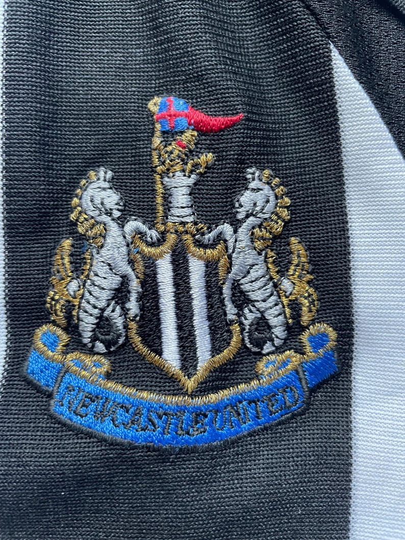 May include: Black and white striped soccer jersey with a black embroidered crest featuring a shield with black and white stripes, two white horses, and a gold crown. The crest is surrounded by a blue ribbon with the text "NEWCASTLE UNITED" in gold.
