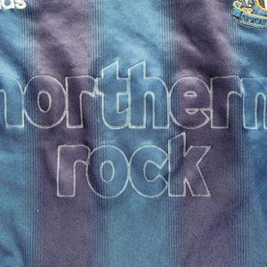 May include: Close-up of a blue and purple striped soccer jersey. The jersey features the text "northern rock" in large white letters. The Adidas logo is visible at the top left, and a team crest is at the top right.