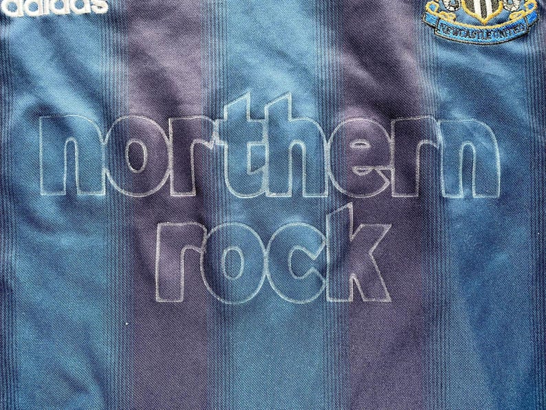 May include: Close-up of a blue and purple striped soccer jersey. The jersey features the text "northern rock" in large white letters. The Adidas logo is visible at the top left, and a team crest is at the top right.