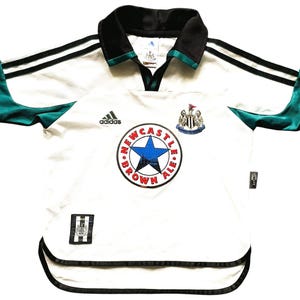 Puede incluir: Camiseta de f&uacute;tbol blanca con detalles en negro y verde azulado. Presenta el logo de Adidas, un escudo de Newcastle Brown Ale y un escudo del equipo. El cuello y las mangas tienen ribetes negros.