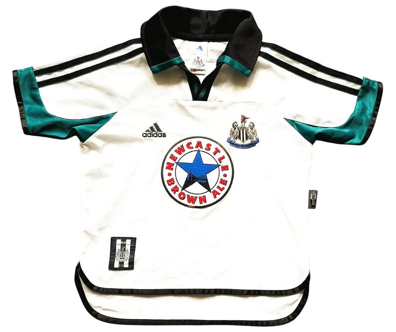 Puede incluir: Camiseta de f&uacute;tbol blanca con detalles en negro y verde azulado. Presenta el logo de Adidas, un escudo de Newcastle Brown Ale y un escudo del equipo. El cuello y las mangas tienen ribetes negros.