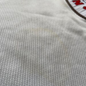 May include: Close-up of a white sports jersey with the Adidas logo at the top. The jersey has a textured pattern and a team logo on the right side. The team logo is circular with red, white, and blue accents.