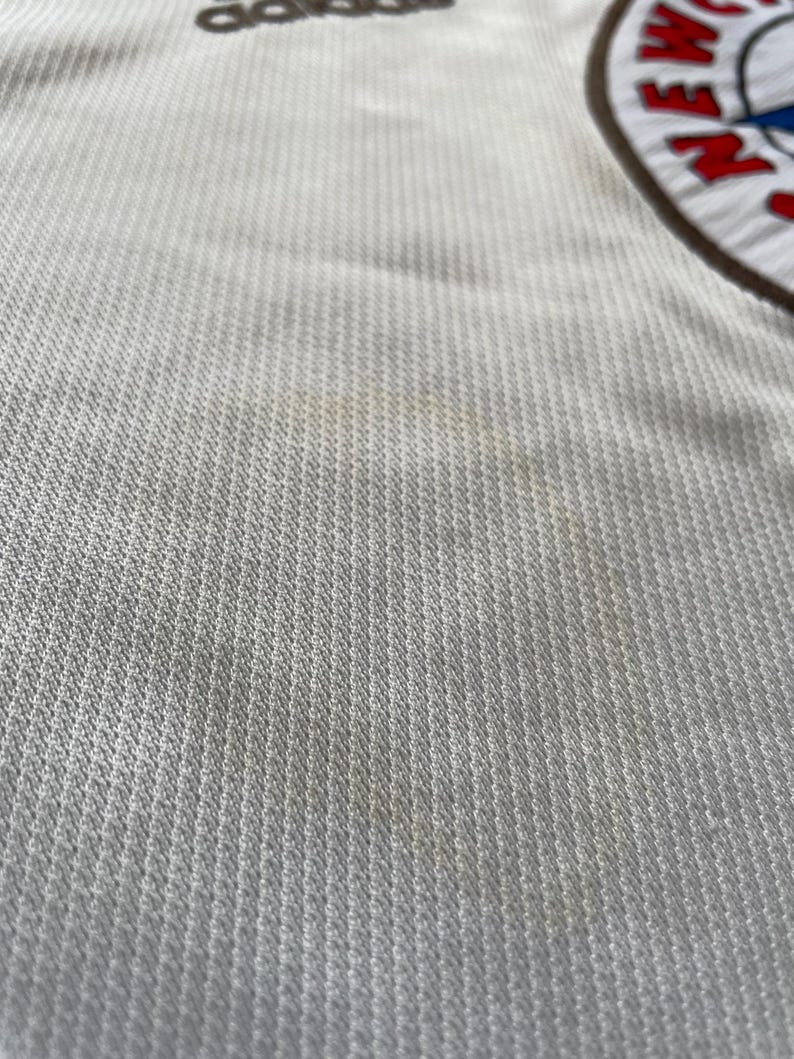 May include: Close-up of a white sports jersey with the Adidas logo at the top. The jersey has a textured pattern and a team logo on the right side. The team logo is circular with red, white, and blue accents.