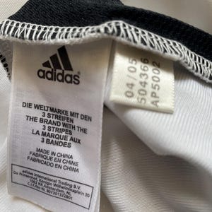 Pode incluir: Close-up de uma etiqueta de roupa branca com o logotipo da Adidas e texto em v&aacute;rios idiomas. A etiqueta inclui as frases "DIE WELTMARKE MIT DEN 3 STREIFEN" e "THE BRAND WITH THE 3 STRIPES". A etiqueta tamb&eacute;m tem uma data e um c&oacute;digo do produto.