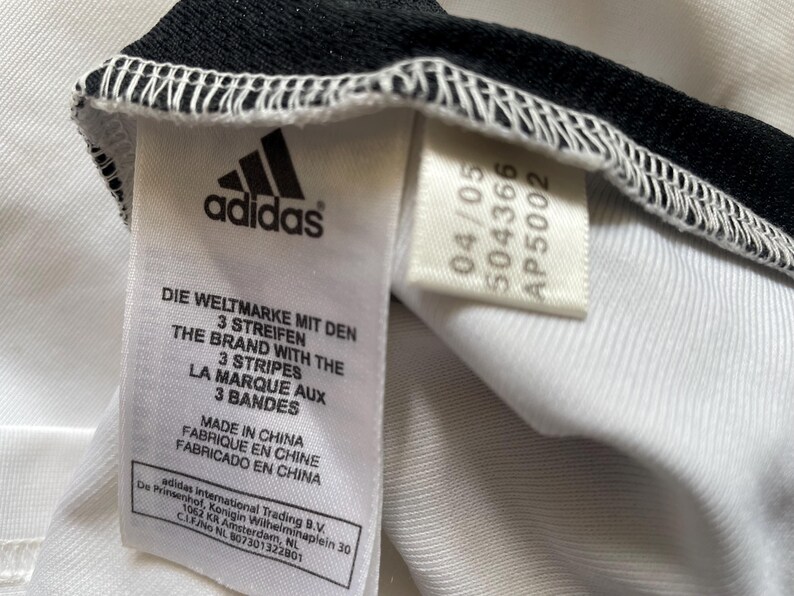 Pode incluir: Close-up de uma etiqueta de roupa branca com o logotipo da Adidas e texto em v&aacute;rios idiomas. A etiqueta inclui as frases "DIE WELTMARKE MIT DEN 3 STREIFEN" e "THE BRAND WITH THE 3 STRIPES". A etiqueta tamb&eacute;m tem uma data e um c&oacute;digo do produto.