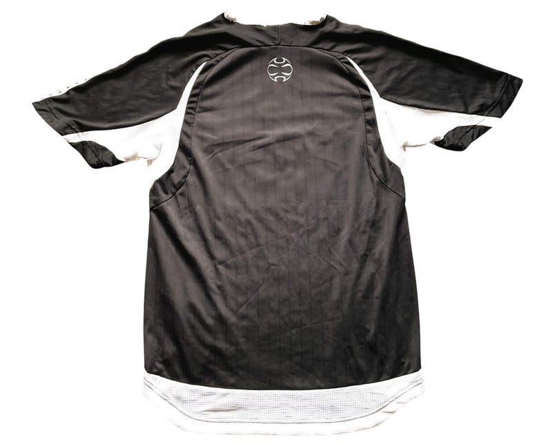 Pu&ograve; includere: Maglia sportiva bianca e nera. La maglia ha maniche corte con pannelli bianchi sui lati e un orlo in rete bianco. Un logo bianco &egrave; sulla parte superiore della schiena. La maglia &egrave; realizzata in materiale atletico traspirante.