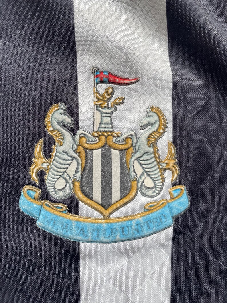 May include: Black and white striped soccer jersey with a crest featuring a shield with a black and white striped design, two seahorses, and the text "NEWCASTLE UNITED".